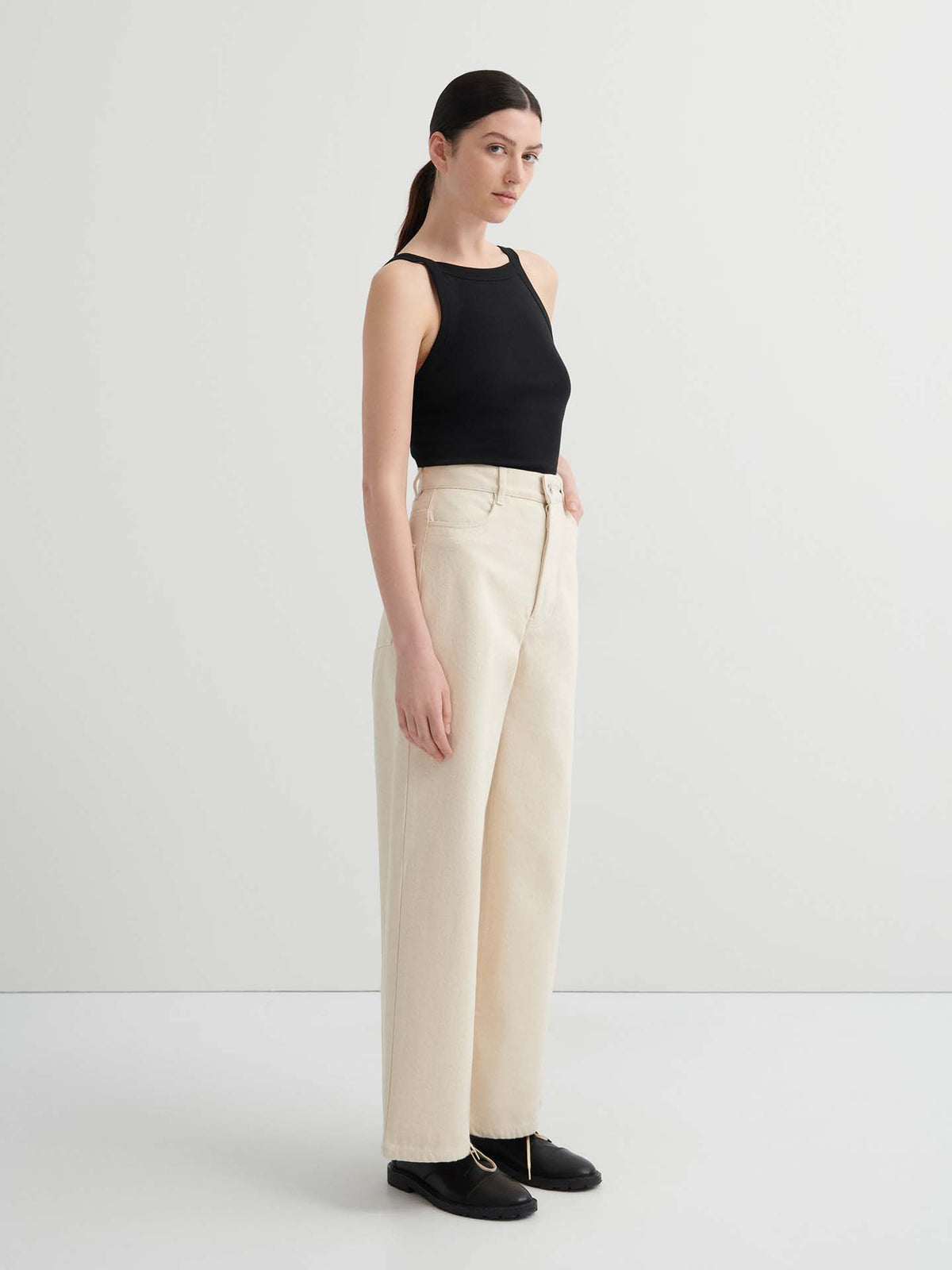 A woman stands in a minimal studio wearing a black sleeveless top, Sailor Jeans – Ecru Denim, and black shoes.