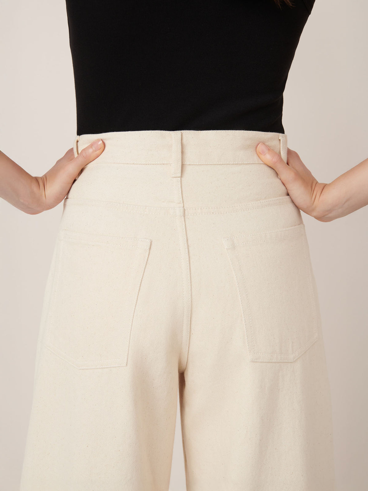A person is shown from the back, hands on hips, wearing Kowtow&#39;s Sailor Jeans in ecru denim paired with a black top.