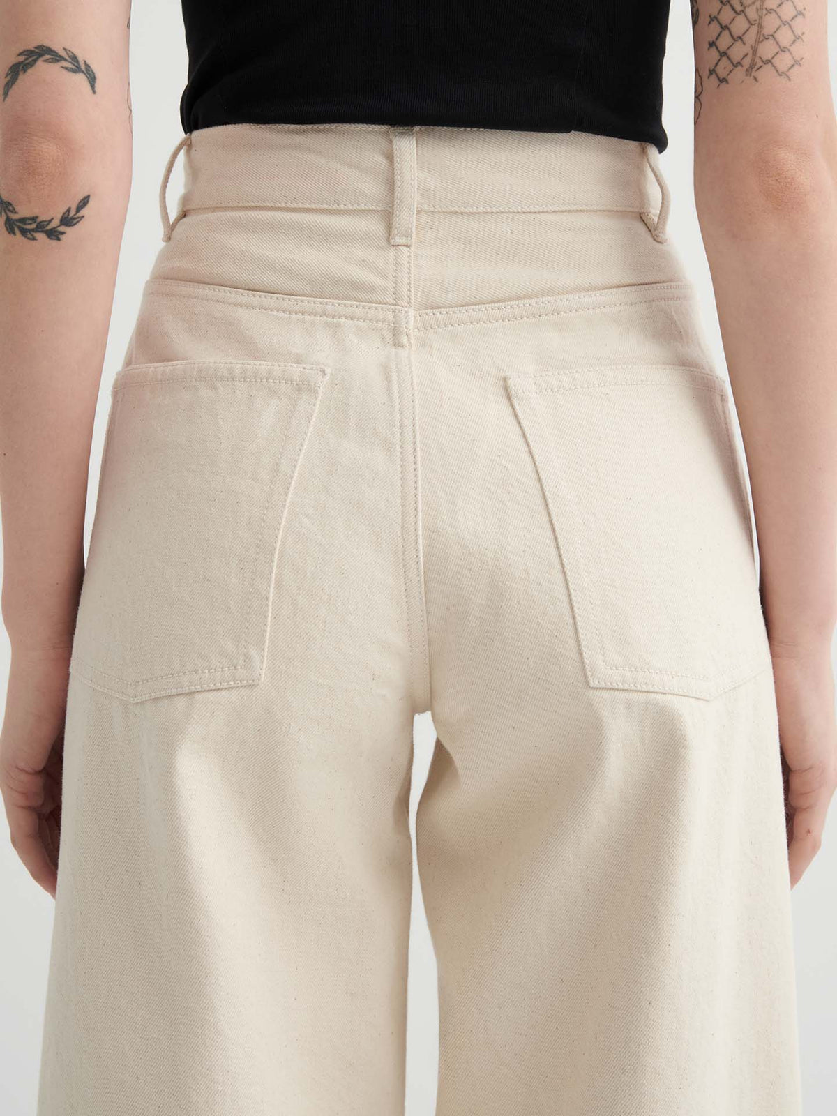 Model wears Sailor Jeans – Ecru Denim, featuring a high-waisted, relaxed fit and large back pockets, paired with a black top. Tattoos on both arms are visible as the look is shown from the back.