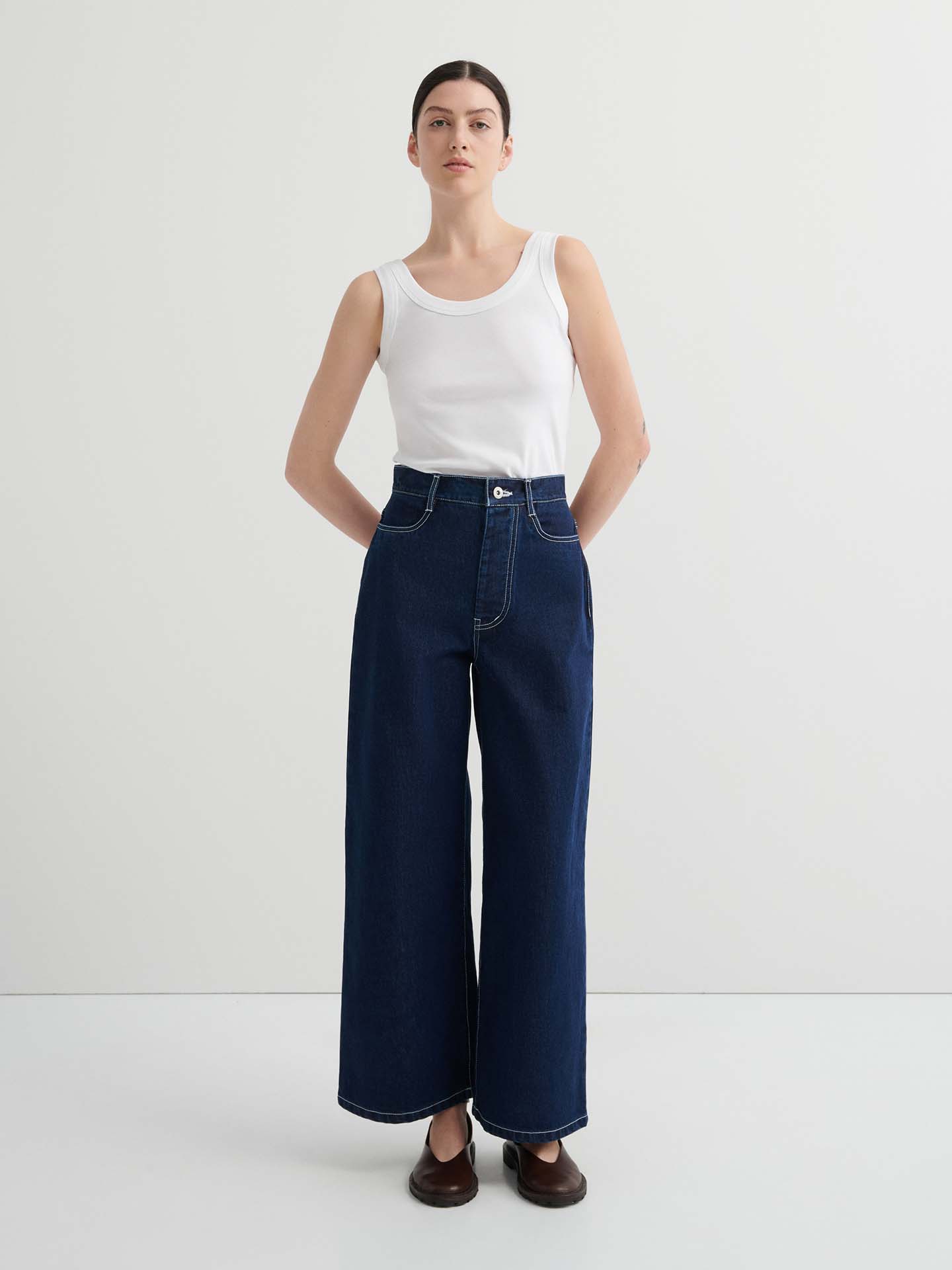 A woman stands against a plain background wearing a white sleeveless top, Sailor Jeans – Indigo, and brown shoes.
