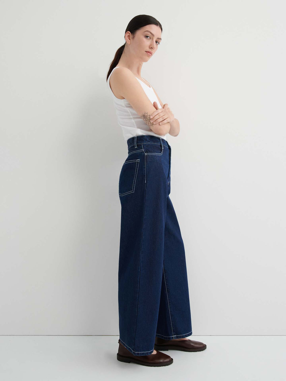 A woman stands against a white wall with arms crossed, wearing a white tank top, Sailor Jeans – Indigo in pre-washed denim, and brown shoes.