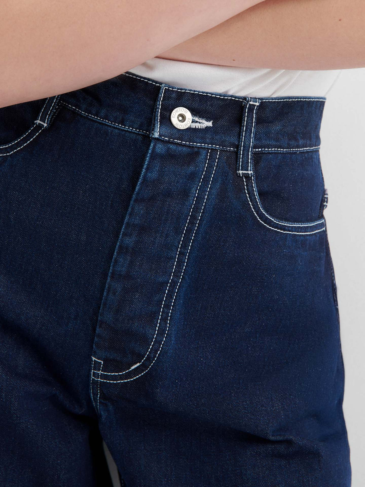 Close-up of a person wearing Sailor Jeans – Indigo, featuring high waist, dark blue pre-washed denim with white stitching, paired with a white top. The image highlights the waistband, button, and front pocket.