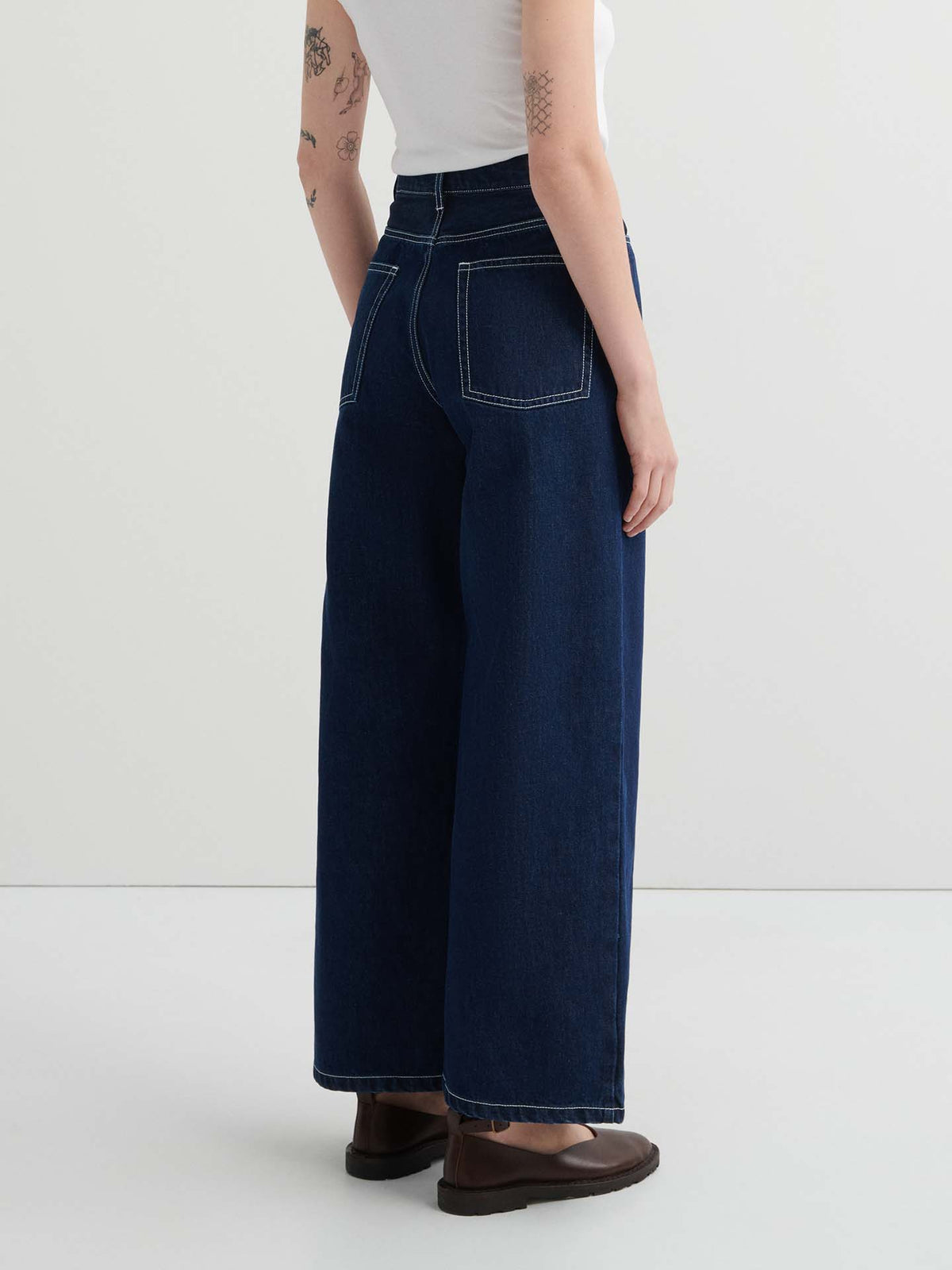 Person wearing a white sleeveless top, Sailor Jeans – Indigo with white stitching, and brown shoes, standing against a plain light background.