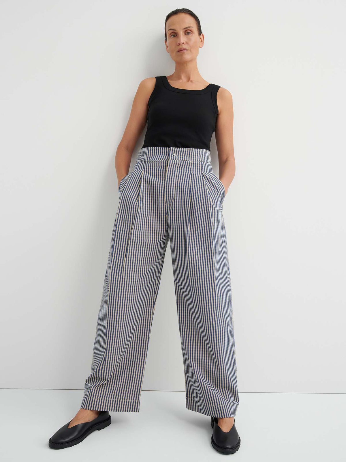 A woman stands against a white wall, hands in her pockets, wearing a black sleeveless top and the Sculpt Pant – Landscape Check: relaxed fit, high-waisted wide-leg plaid trousers with a fixed waistband, paired with black shoes.