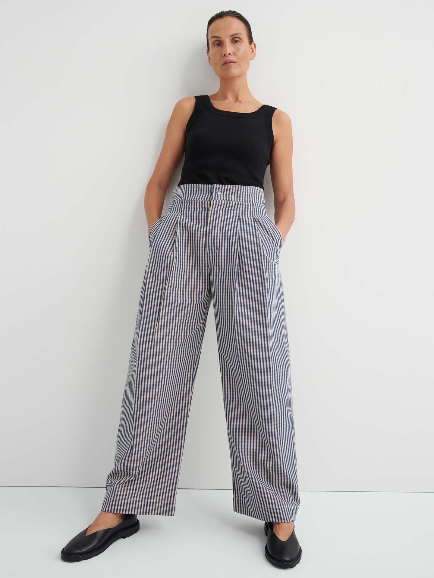 A woman stands against a white wall, hands in her pockets, wearing a black sleeveless top and the Sculpt Pant – Landscape Check: relaxed fit, high-waisted wide-leg plaid trousers with a fixed waistband, paired with black shoes.