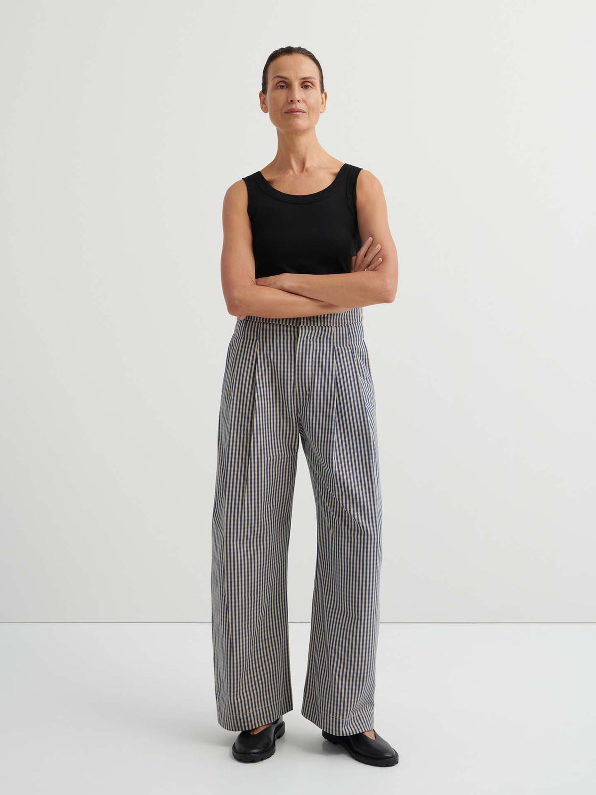 A woman stands against a plain background wearing a sleeveless black top and high-waisted Sculpt Pant – Landscape Check with a relaxed fit and fixed waistband. Her arms are crossed, and black shoes complete the look.