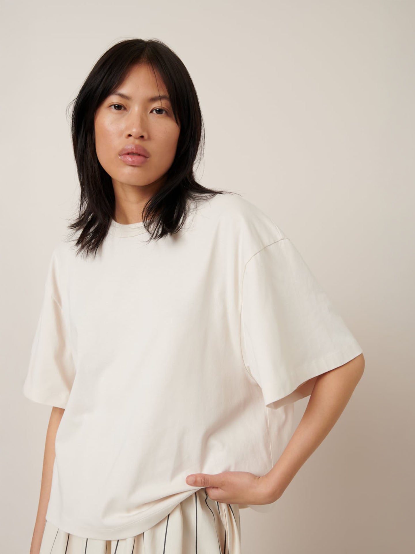 A person with long dark hair wears Kowtow's Shadow Tee in Whisper—an oversized white T-shirt—and striped pants, standing against a neutral background.