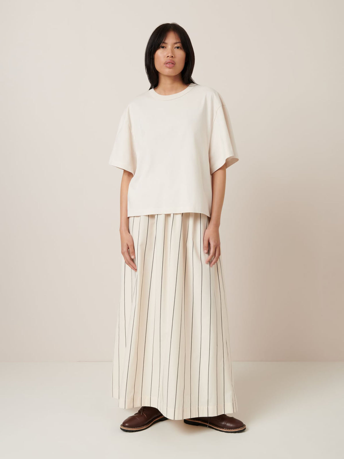 A person wearing the Kowtow Shadow Tee in Whisper stands against a plain background, showcasing an oversized fit paired with a long pleated skirt featuring bold vertical stripes and brown shoes.