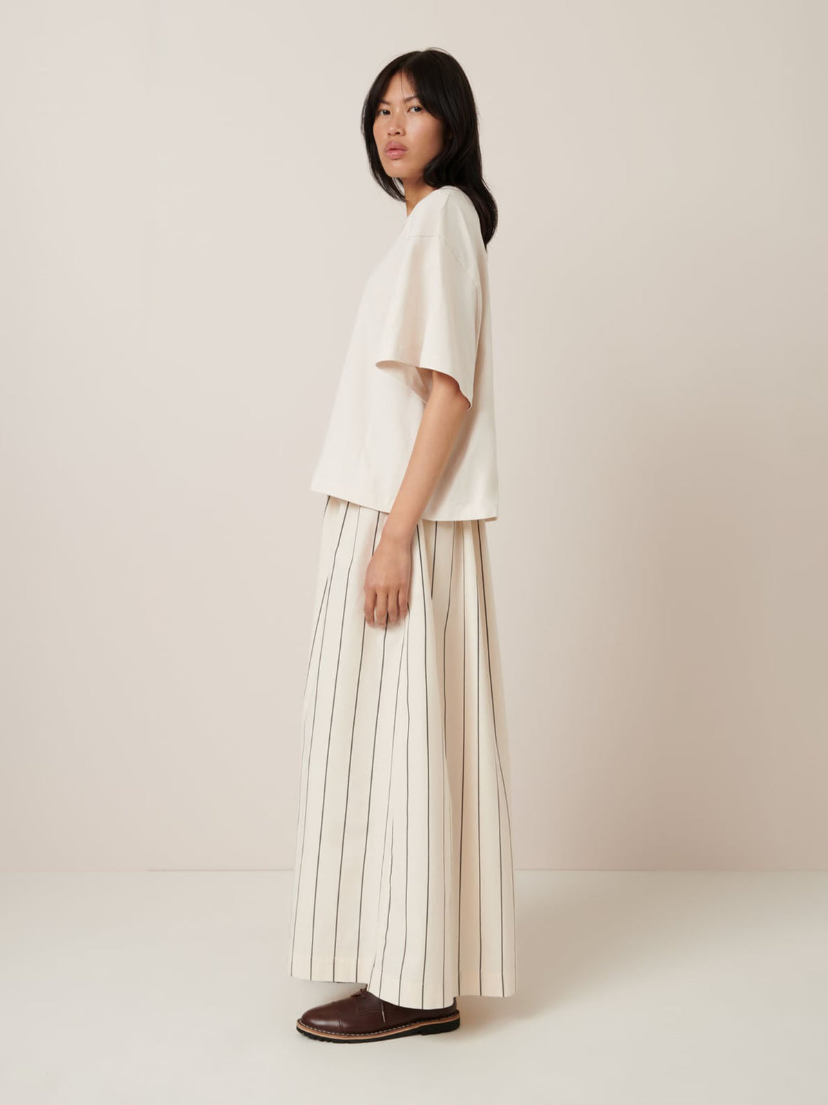 A person stands sideways in an oversized fit, wearing Kowtow&#39;s Shadow Tee – Whisper, a long white skirt with vertical black stripes highlighting the garment pattern, and brown shoes against a plain background.