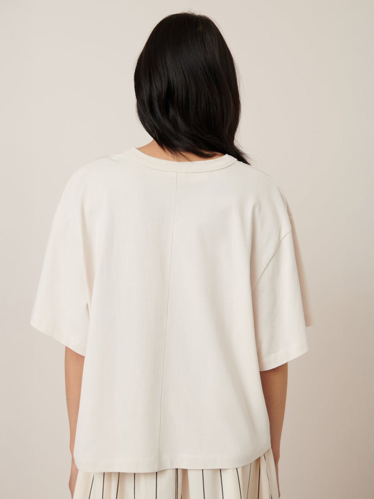 The person, embodying effortless style, stands facing away against a neutral backdrop with long black hair cascading down the back of a Kowtow oversized fit white Shadow Tee – Whisper.