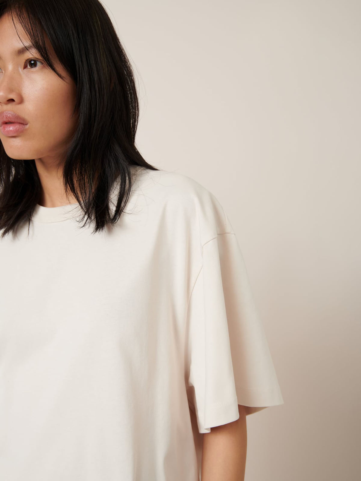 A person with long black hair is wearing the Kowtow Shadow Tee – Whisper, featuring an oversized fit, in front of a beige background.