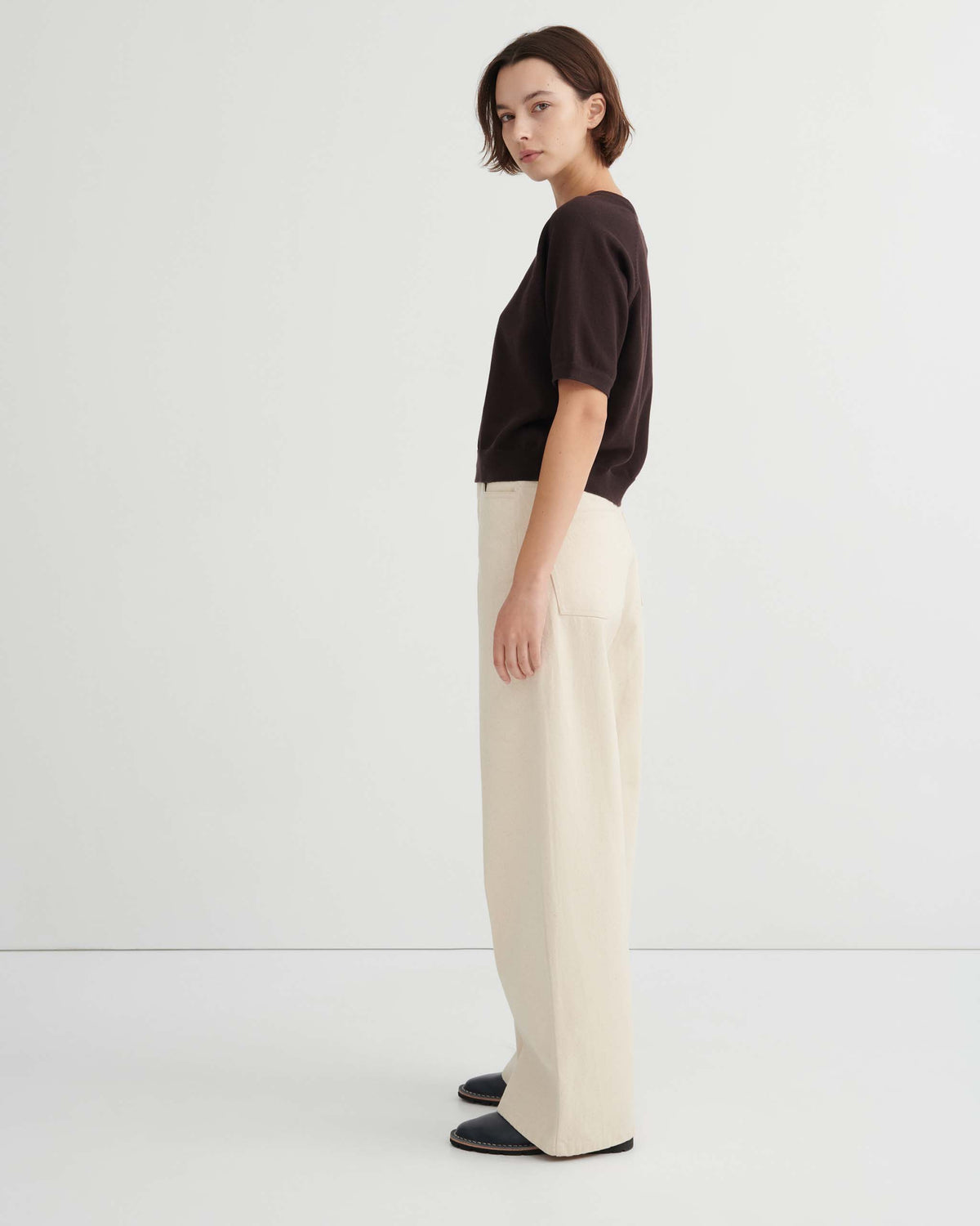 Person standing in profile against a plain white wall, wearing a dark short-sleeve top, cream wide-leg pants, and black shoes.