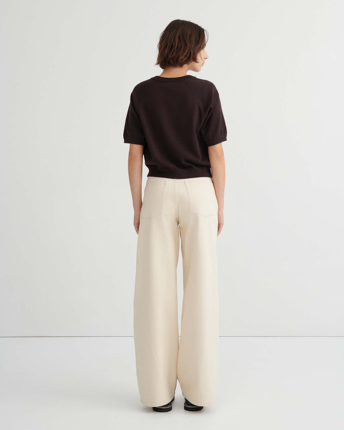 A person stands facing away, wearing a black short-sleeve top and high-waisted, wide-leg cream pants, against a plain white background.