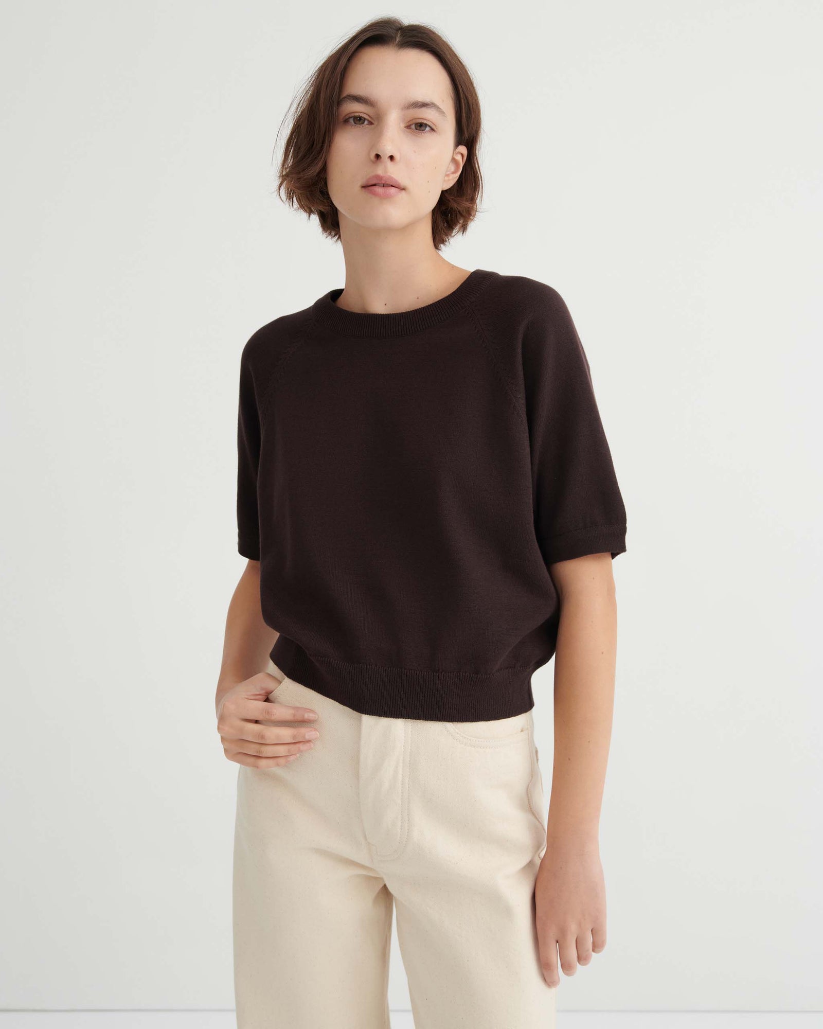 A person with short brown hair wears a dark short-sleeved sweater and light pants, standing against a plain white background.