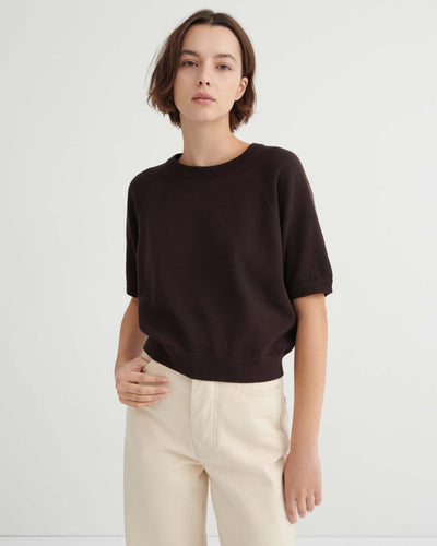 A person with short brown hair wears a dark short-sleeved sweater and light pants, standing against a plain white background.