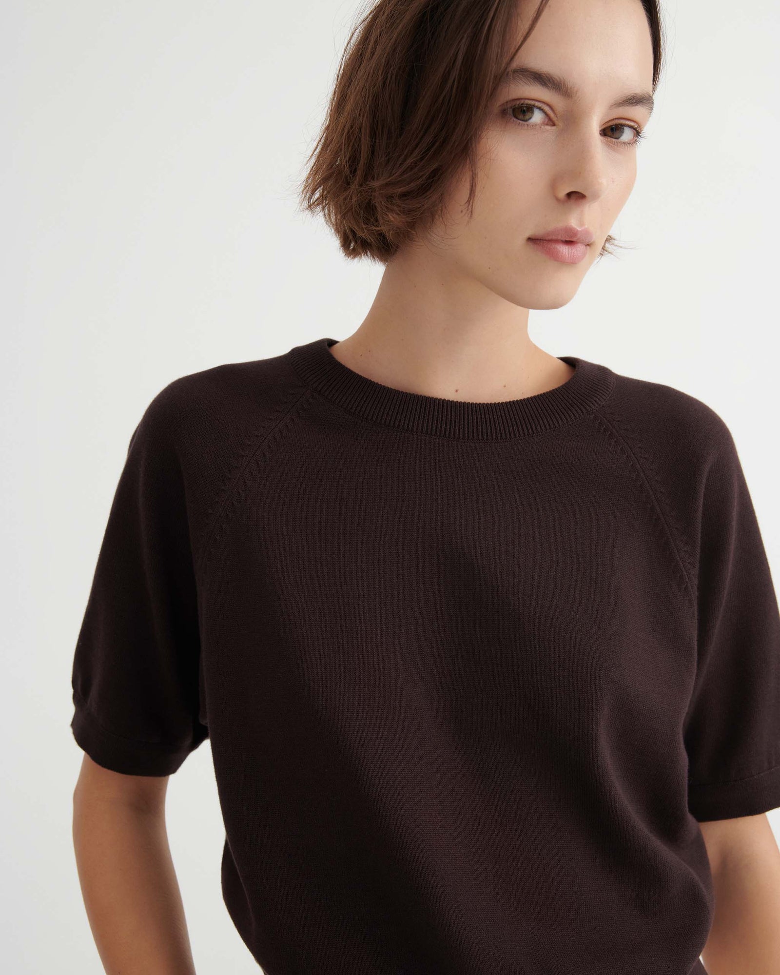 A person with short brown hair wears a dark short-sleeved sweater and light pants, standing against a plain white background.