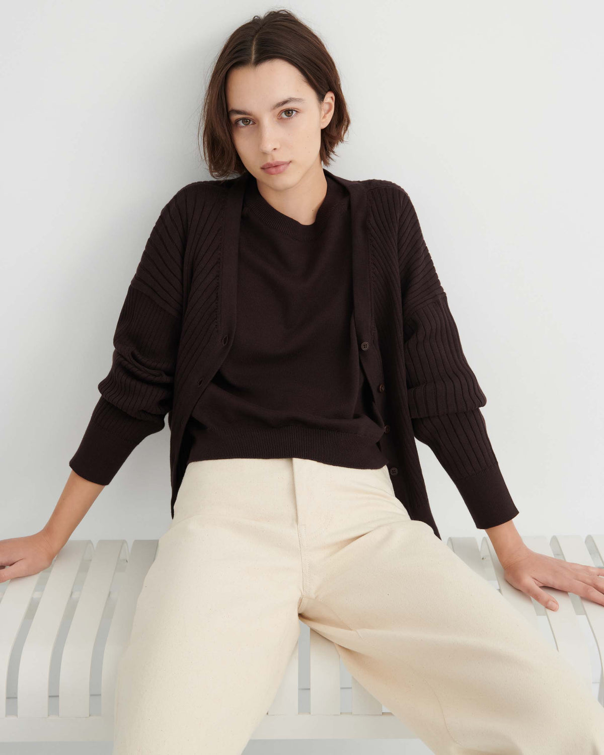 Woman with straight brown hair wearing a black sweater and cardigan with cream wide-leg pants sits on a white bench against a plain white wall.