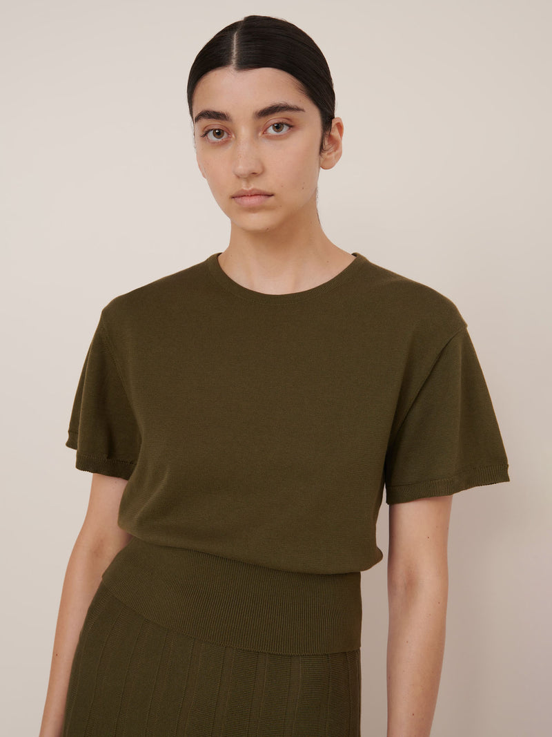 A woman with dark hair tied back wears the Kowtow Silhouette Top – Ivy, styled with a matching ribbed skirt, against a plain light background. This olive green top offers a standard fit that's true to size for everyday versatility.