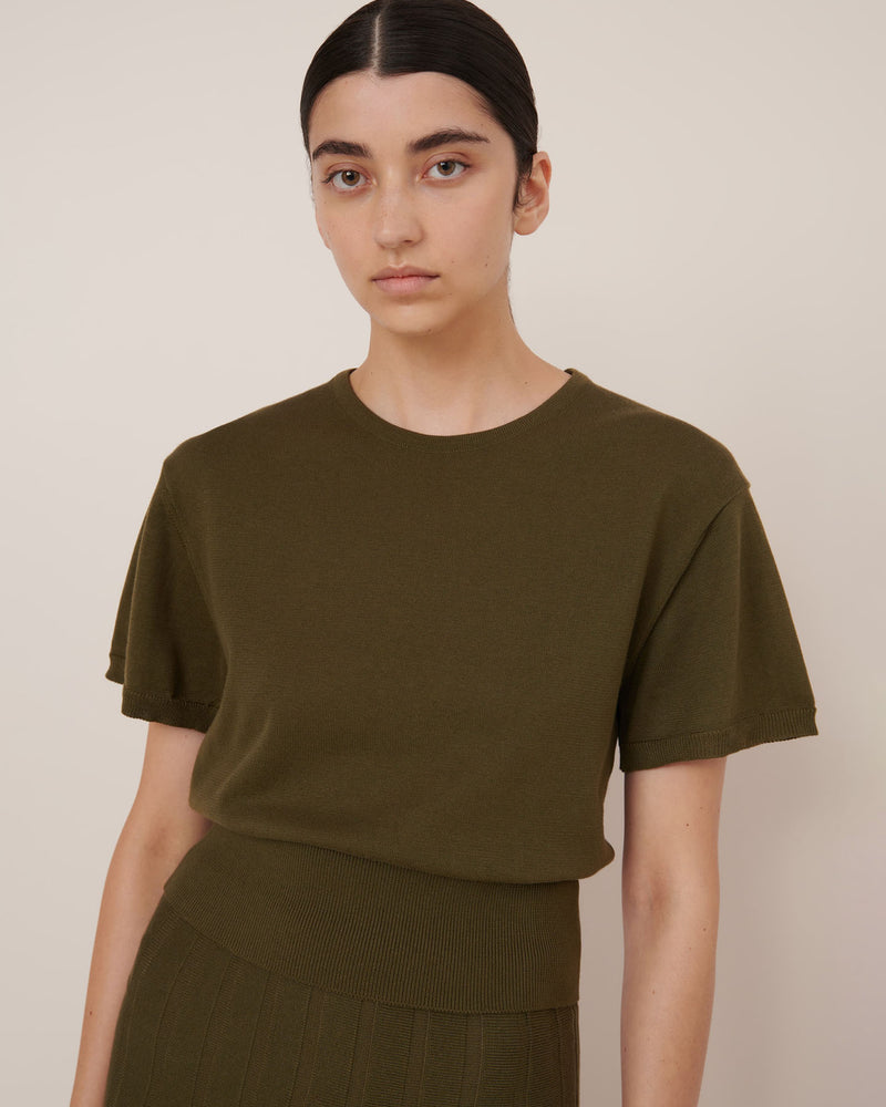 A woman with dark hair tied back wears the Kowtow Silhouette Top – Ivy, styled with a matching ribbed skirt, against a plain light background. This olive green top offers a standard fit that's true to size for everyday versatility.