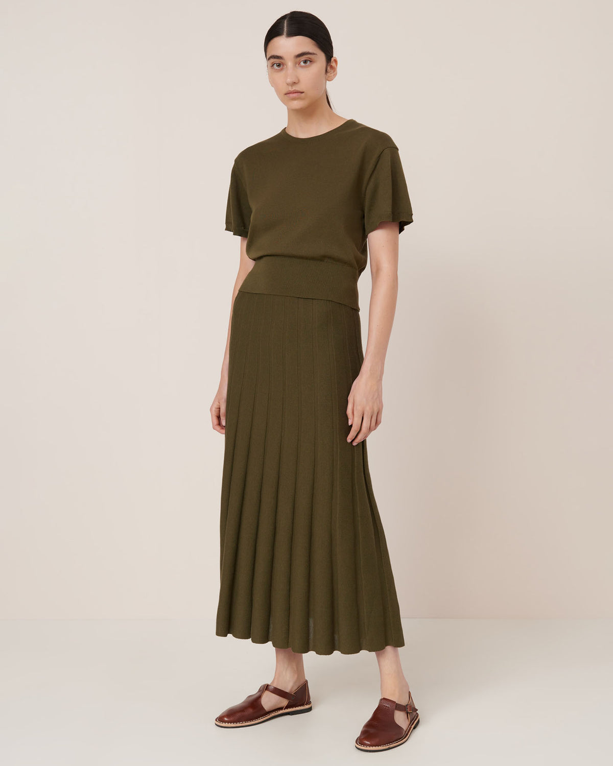 A person stands against a plain background wearing the Kowtow Silhouette Top – Ivy and matching pleated skirt with brown sandals, styled in a standard fit that’s true to size.