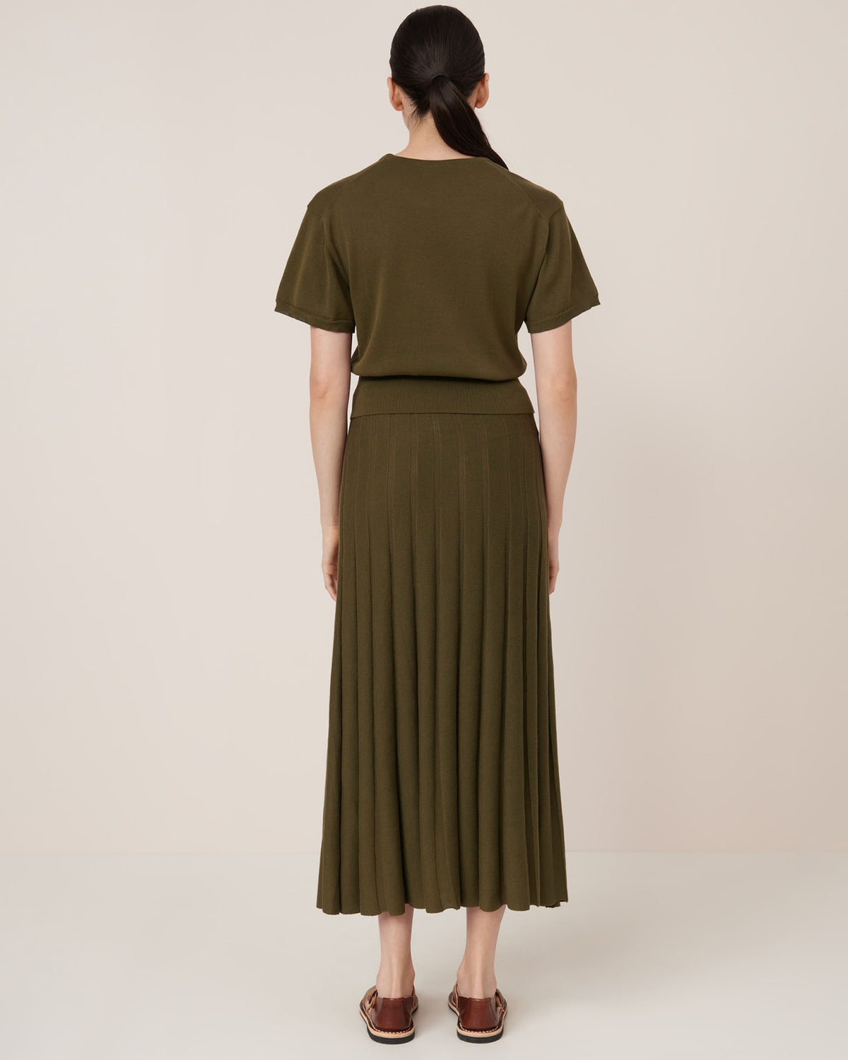 A woman faces away, wearing the Kowtow Silhouette Top in Ivy and a matching pleated midi skirt with brown flat sandals, against a light background—styled in a standard fit that's true to size.