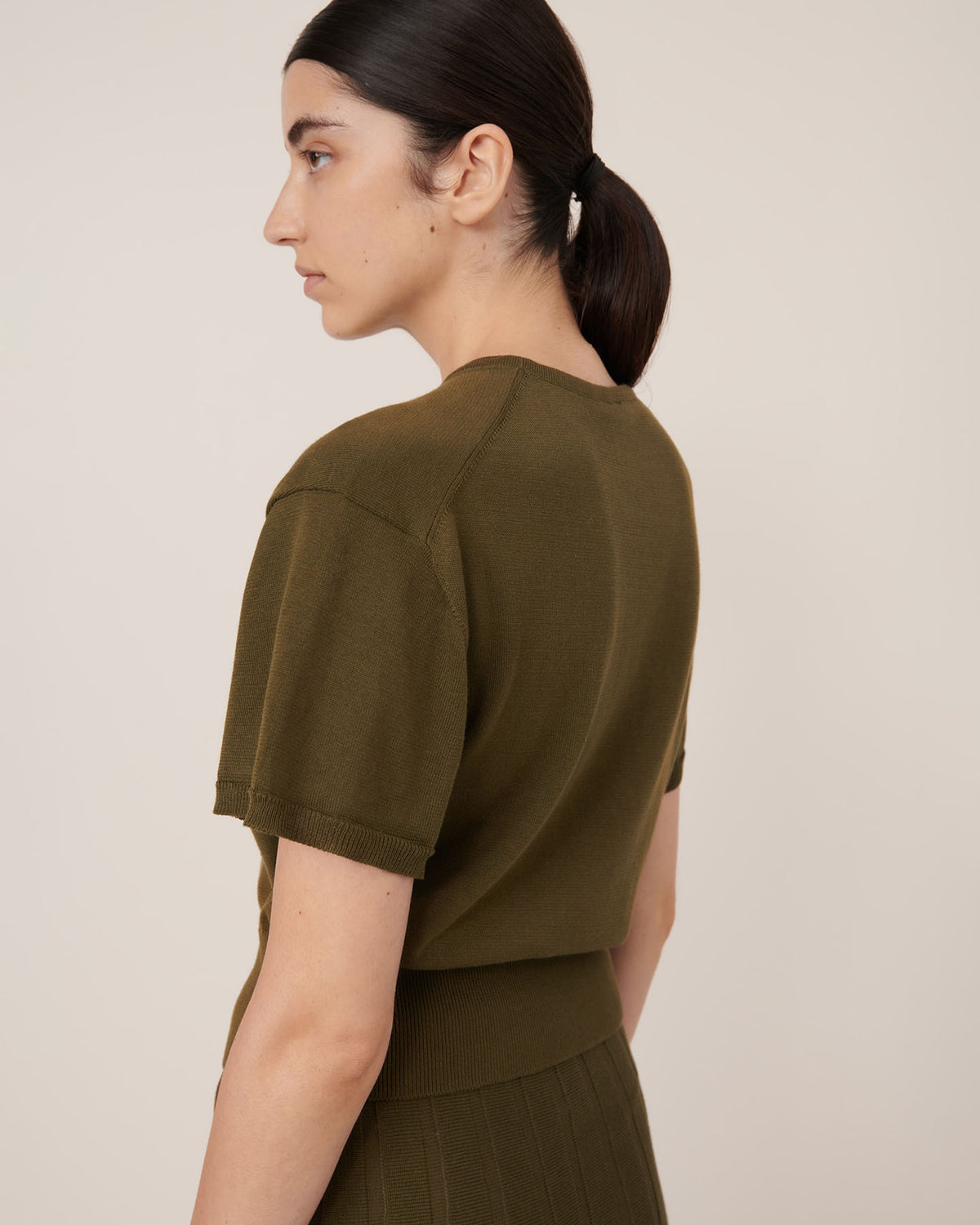 A person with dark hair in a low ponytail wears the Kowtow Silhouette Top – Ivy, an olive green short-sleeve knit, shown from the side against a neutral background; fit is standard and true to size.