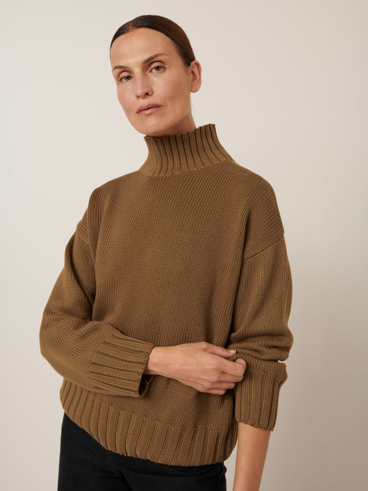 Person with straight brown hair in a low bun wears the Kowtow Staple Sweater – Dark Spice, a relaxed-fit ribbed turtleneck, with black pants against a plain light background. Check the size guide for garment measurements.