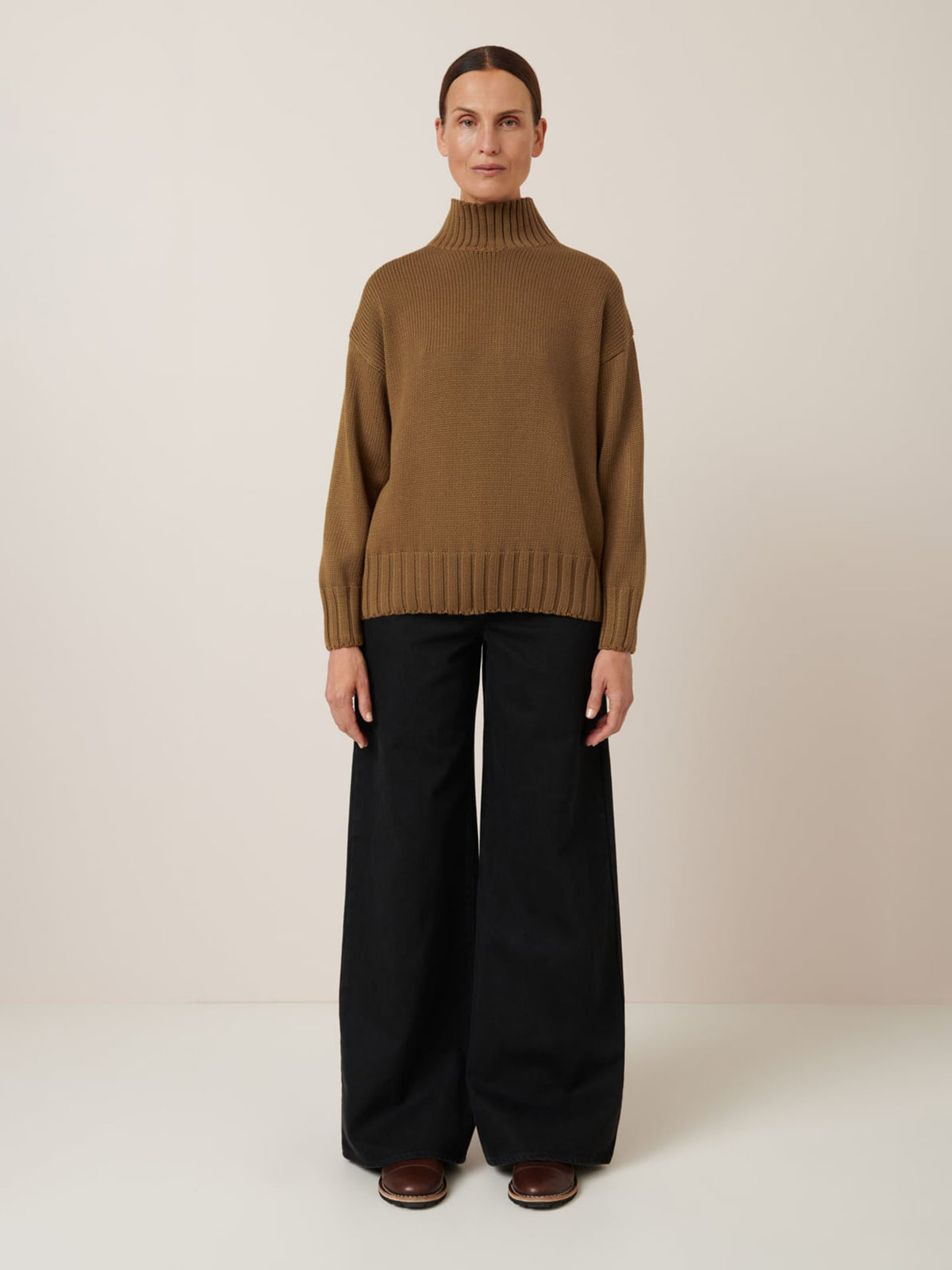 A person faces forward, wearing the Kowtow Staple Sweater in Dark Spice with wide-leg black pants and brown shoes, set against a plain light background.