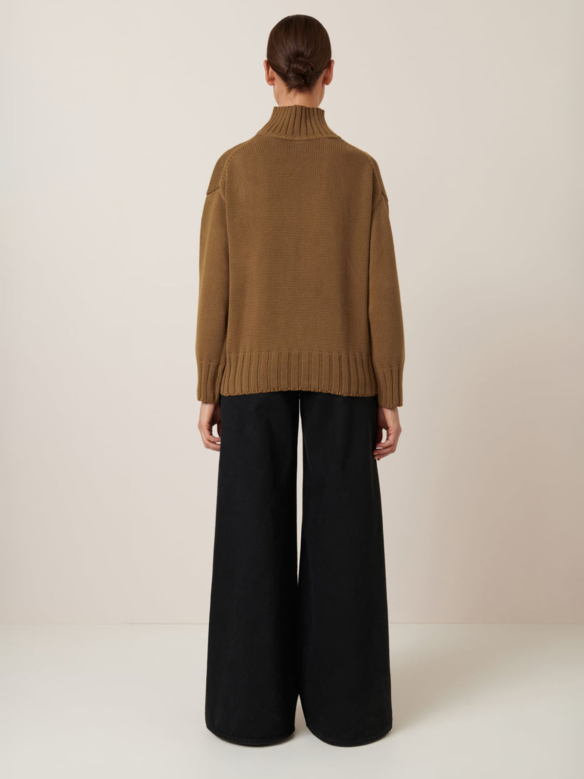 A person stands facing away from the camera, wearing Kowtow’s Staple Sweater in Dark Spice and wide-leg black pants against a plain off-white background. Refer to our size guide for precise garment measurements.