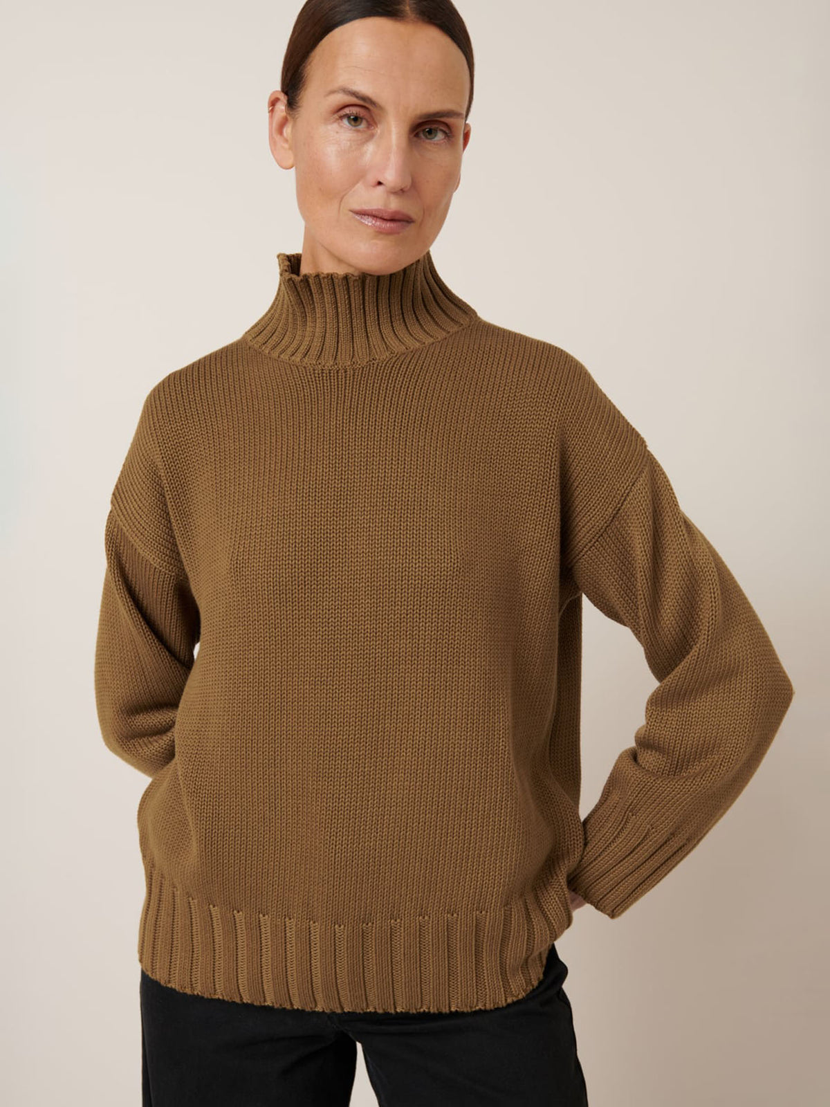 A woman with brown hair in a low bun wears the Kowtow Staple Sweater in Dark Spice and black pants, standing against a plain light background.