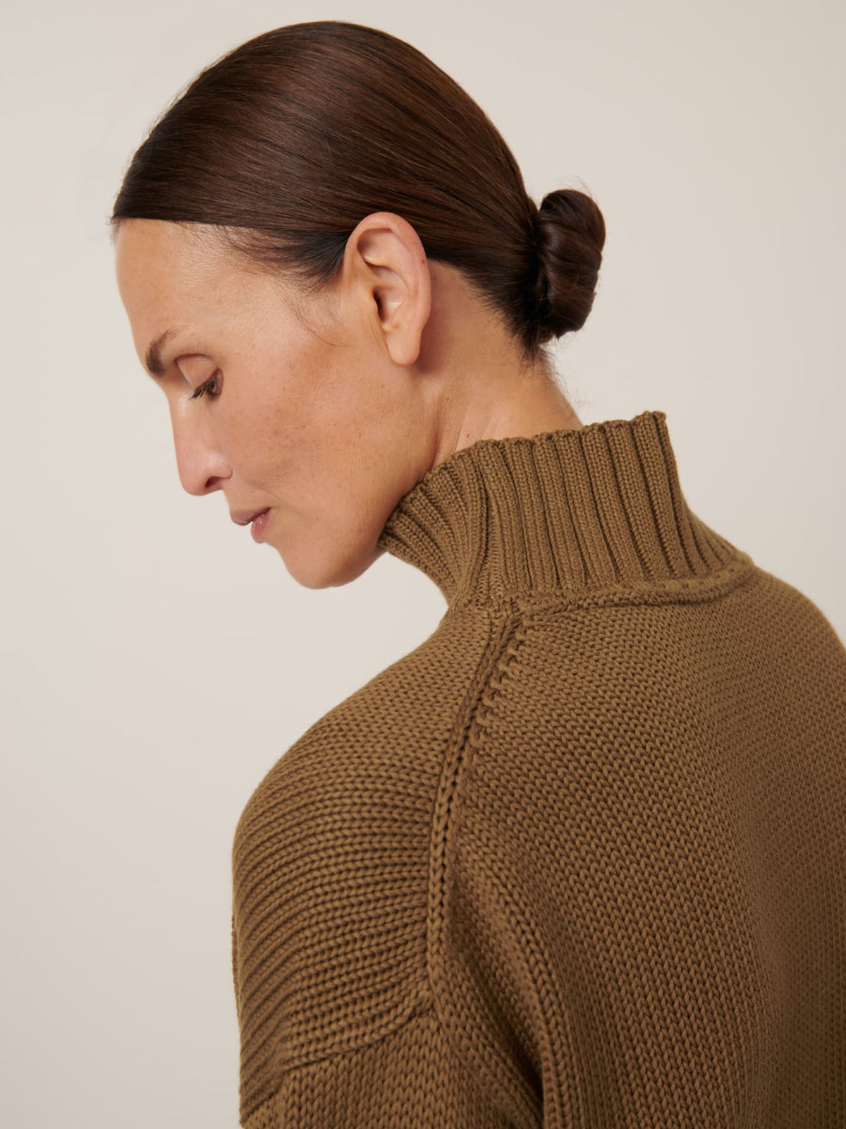 A person with dark hair in a low bun wears the Kowtow Staple Sweater in Dark Spice, a relaxed-fit ribbed turtleneck, shown in profile against a plain background.