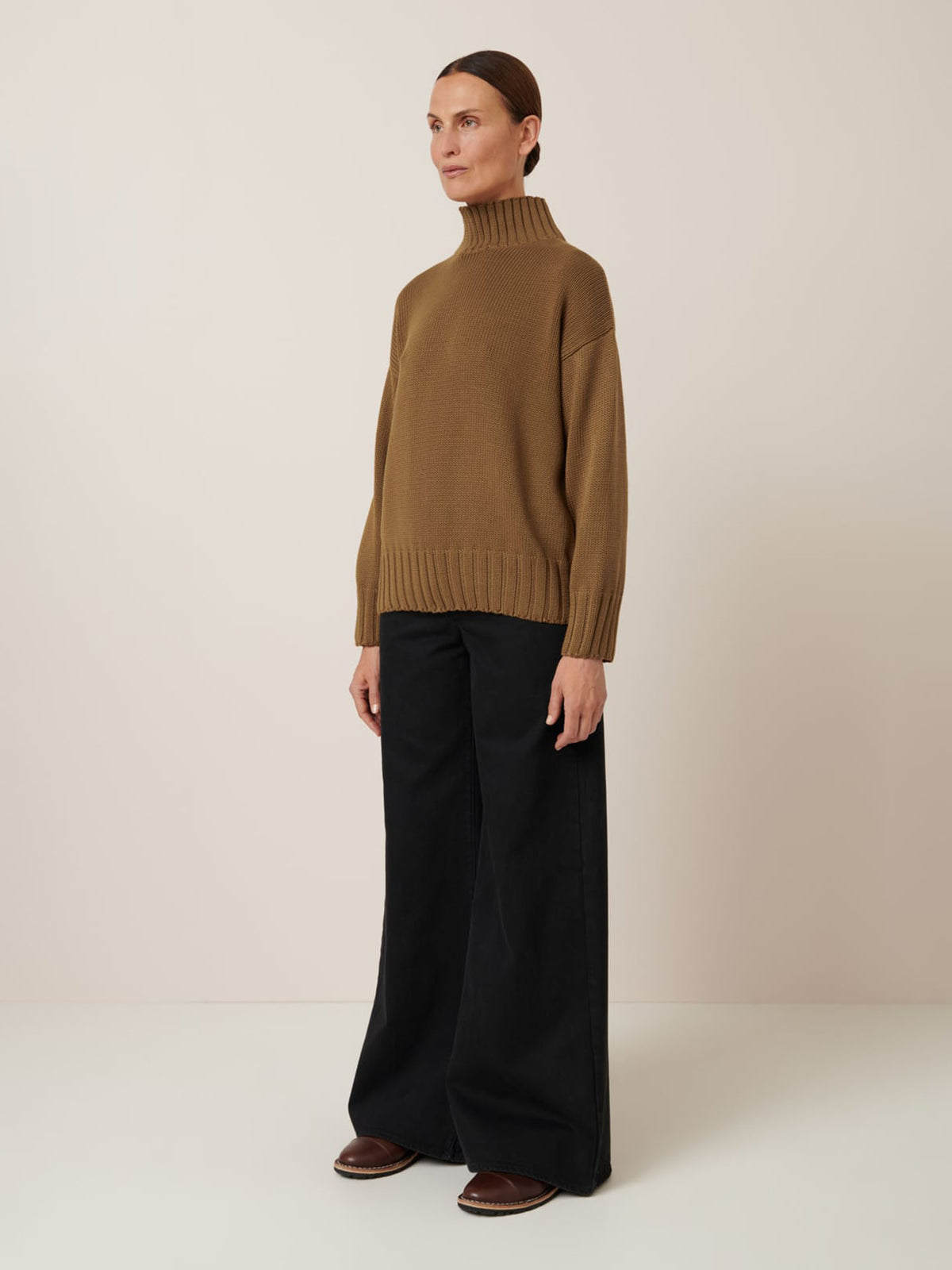 A woman stands against a plain background wearing the Kowtow Staple Sweater in Dark Spice, paired with wide-leg black pants and brown shoes.