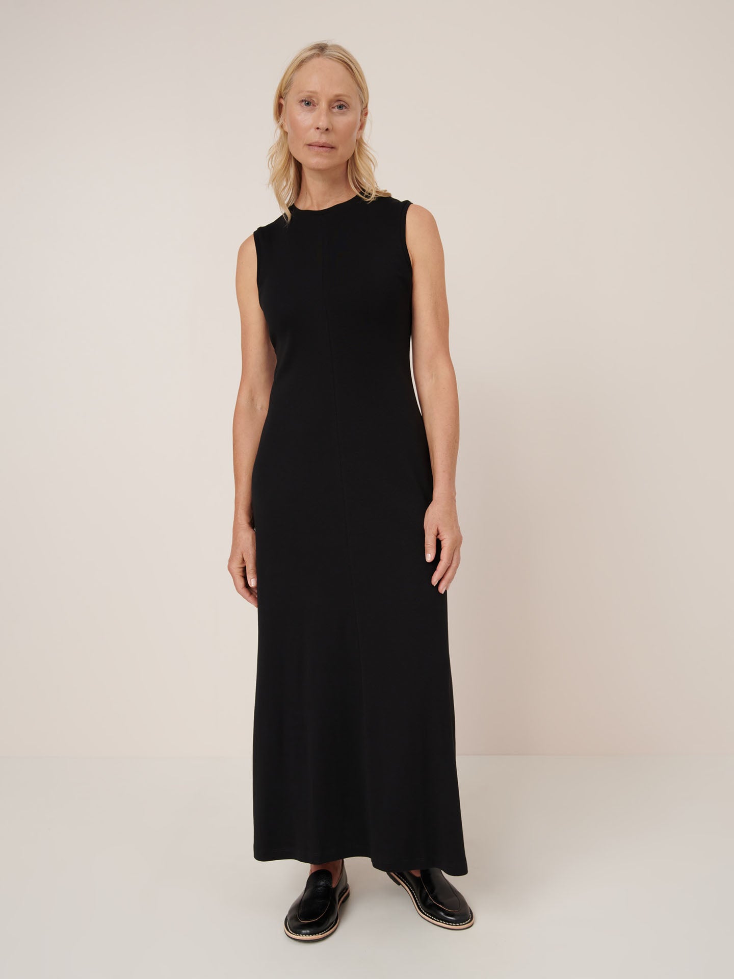 A woman poses against a plain background wearing the Kowtow Still Life Dress – Black, a close-fit, sleeveless, ankle-length dress styled with black shoes.