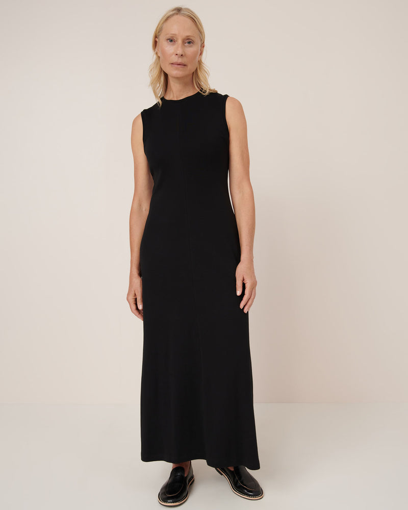 A woman poses against a plain background wearing the Kowtow Still Life Dress – Black, a close-fit, sleeveless, ankle-length dress styled with black shoes.