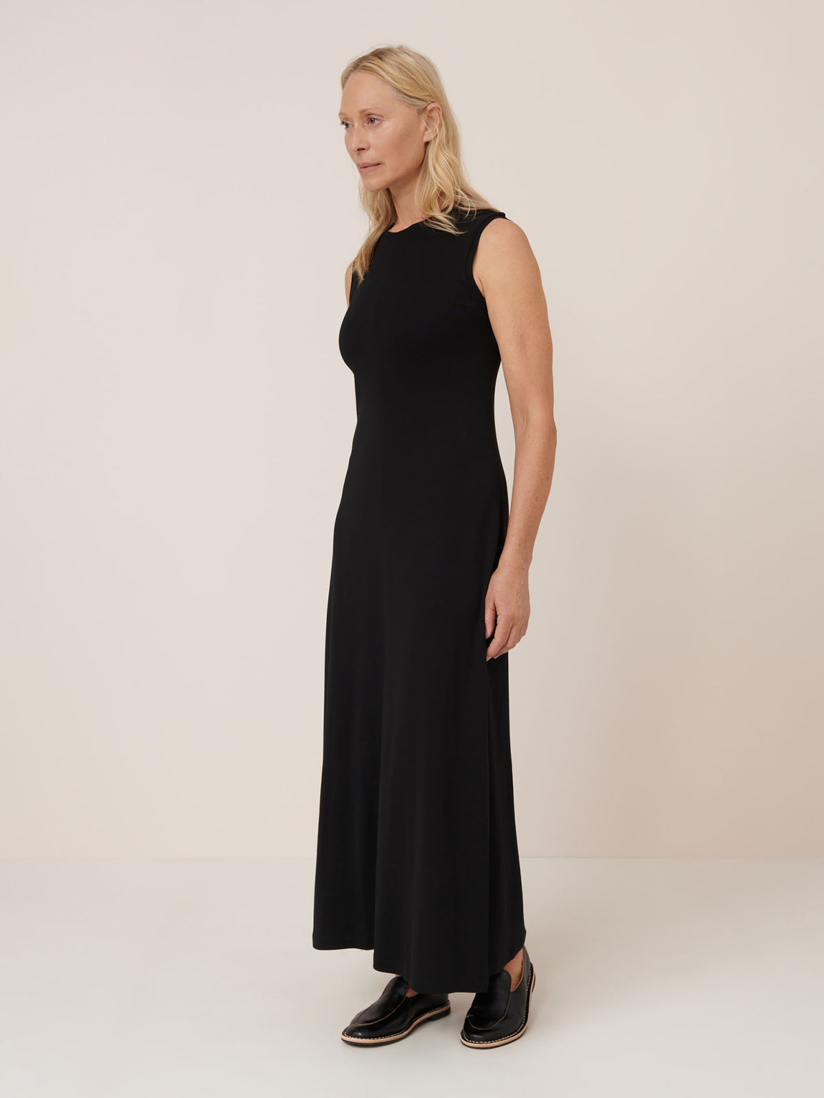 A woman with blonde hair wears the Kowtow Still Life Dress in black—a sleeveless, ankle-length, close-fitting dress—paired with black slip-on shoes, standing against a plain light background.