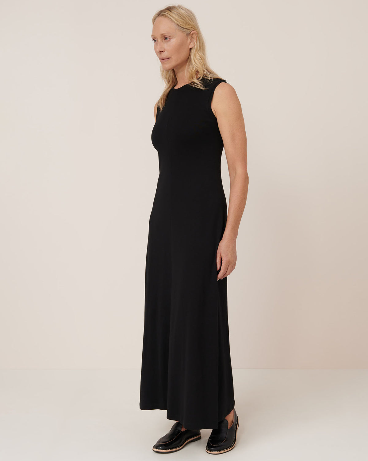 A woman with blonde hair wears the Kowtow Still Life Dress in black—a sleeveless, ankle-length, close-fitting dress—paired with black slip-on shoes, standing against a plain light background.