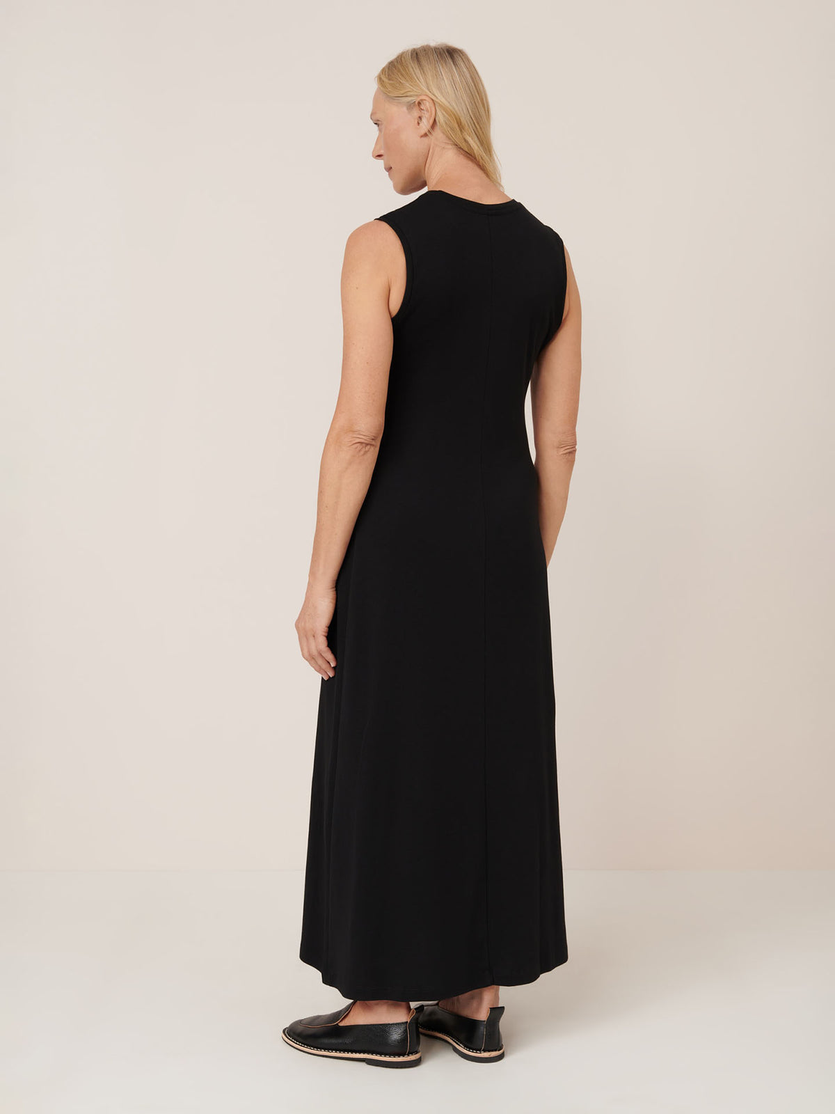 A woman faces away, wearing the Kowtow Still Life Dress – Black, a sleeveless, ankle-length, close-fit dress with black slip-on shoes against a plain light background. Check the size guide for exact measurements.