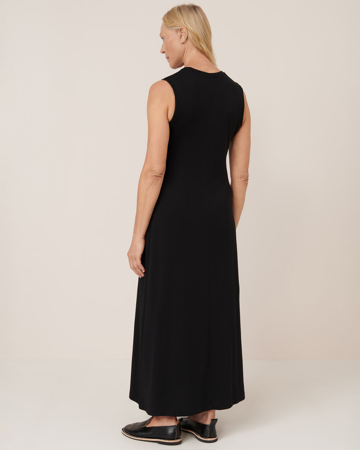 A woman faces away, wearing the Kowtow Still Life Dress – Black, a sleeveless, ankle-length, close-fit dress with black slip-on shoes against a plain light background. Check the size guide for exact measurements.