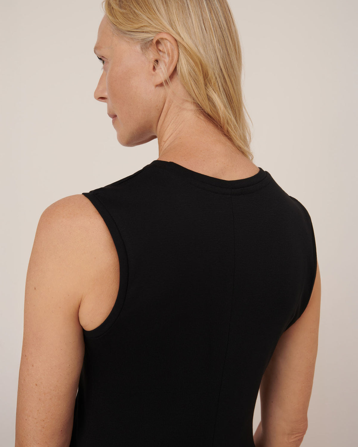 A person with light skin and blonde hair is shown from the back, wearing the Kowtow Still Life Dress in black, a sleeveless close-fitting piece, set against a plain background.