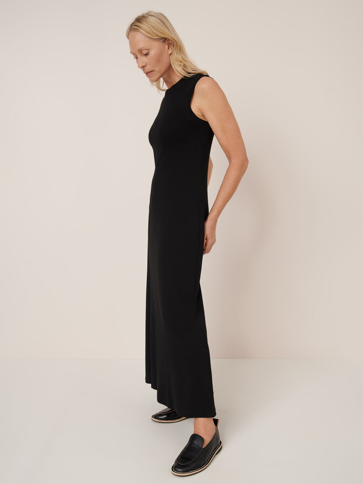 A person in profile wears the Kowtow Still Life Dress – Black, a sleeveless, ankle-length, close-fitting black dress, paired with black slip-on shoes against a plain background.