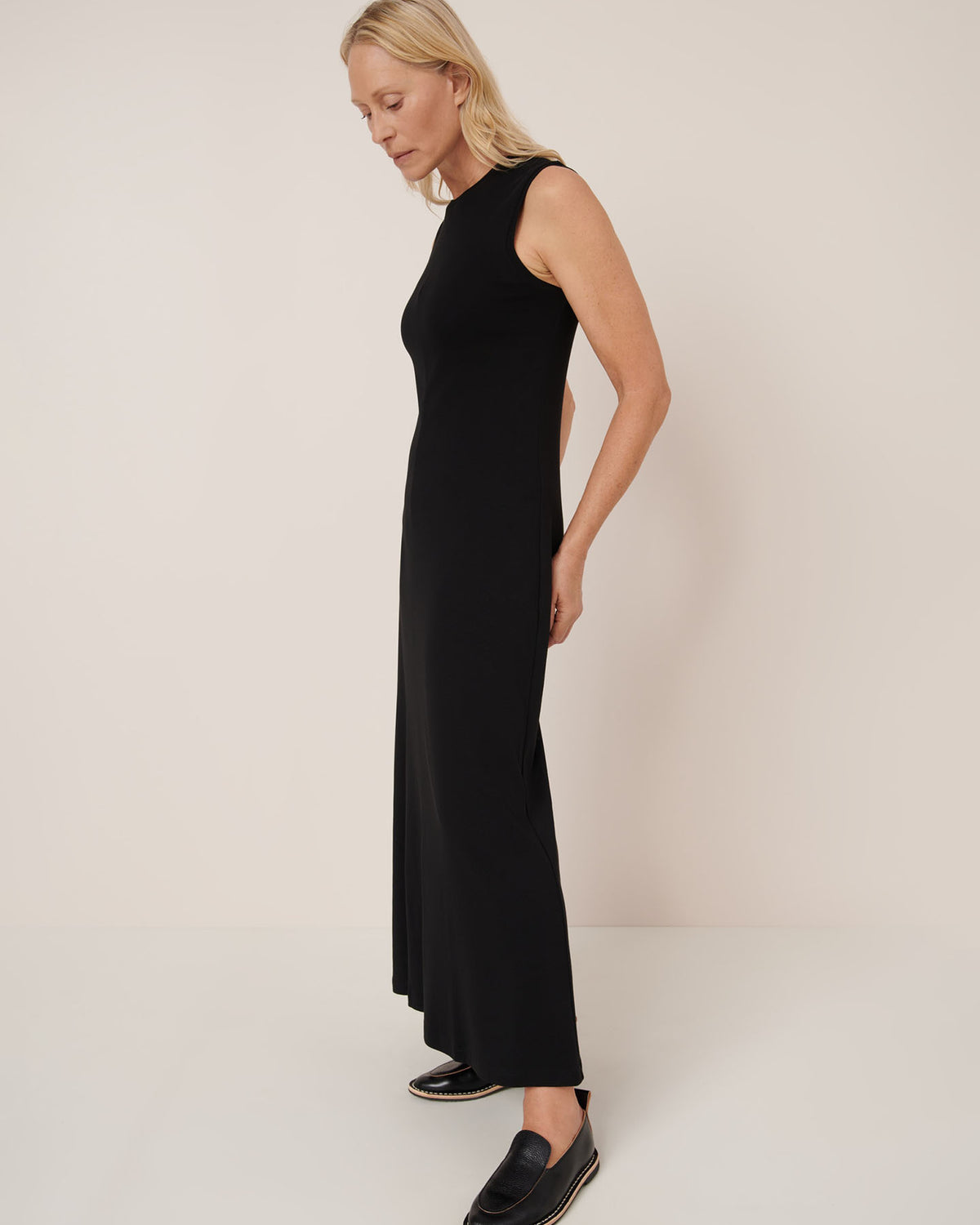 A person in profile wears the Kowtow Still Life Dress – Black, a sleeveless, ankle-length, close-fitting black dress, paired with black slip-on shoes against a plain background.
