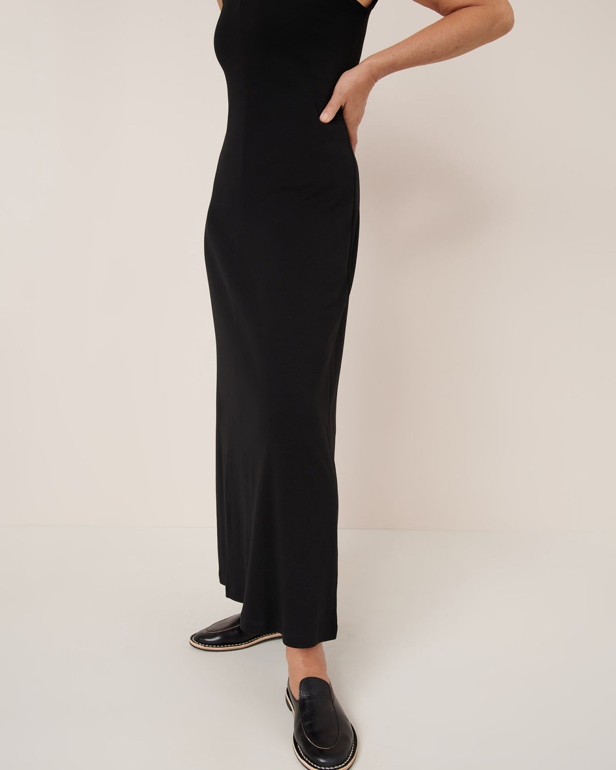 A person wears the Kowtow Still Life Dress – Black, a sleeveless fitted maxi, paired with black slip-on shoes, standing against a plain light background.