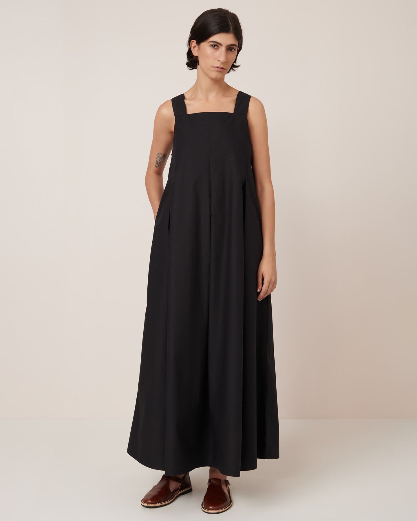 A person with short dark hair wears the Kowtow Study Dress in black, a sleeveless, loose-fitting maxi dress, paired with brown slip-on shoes and stands against a plain light background.