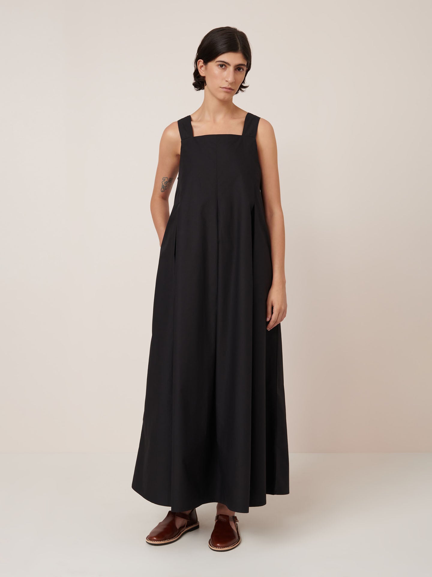 A person with short dark hair wears the Kowtow Study Dress in black, a sleeveless, loose-fitting maxi dress, paired with brown slip-on shoes and stands against a plain light background.