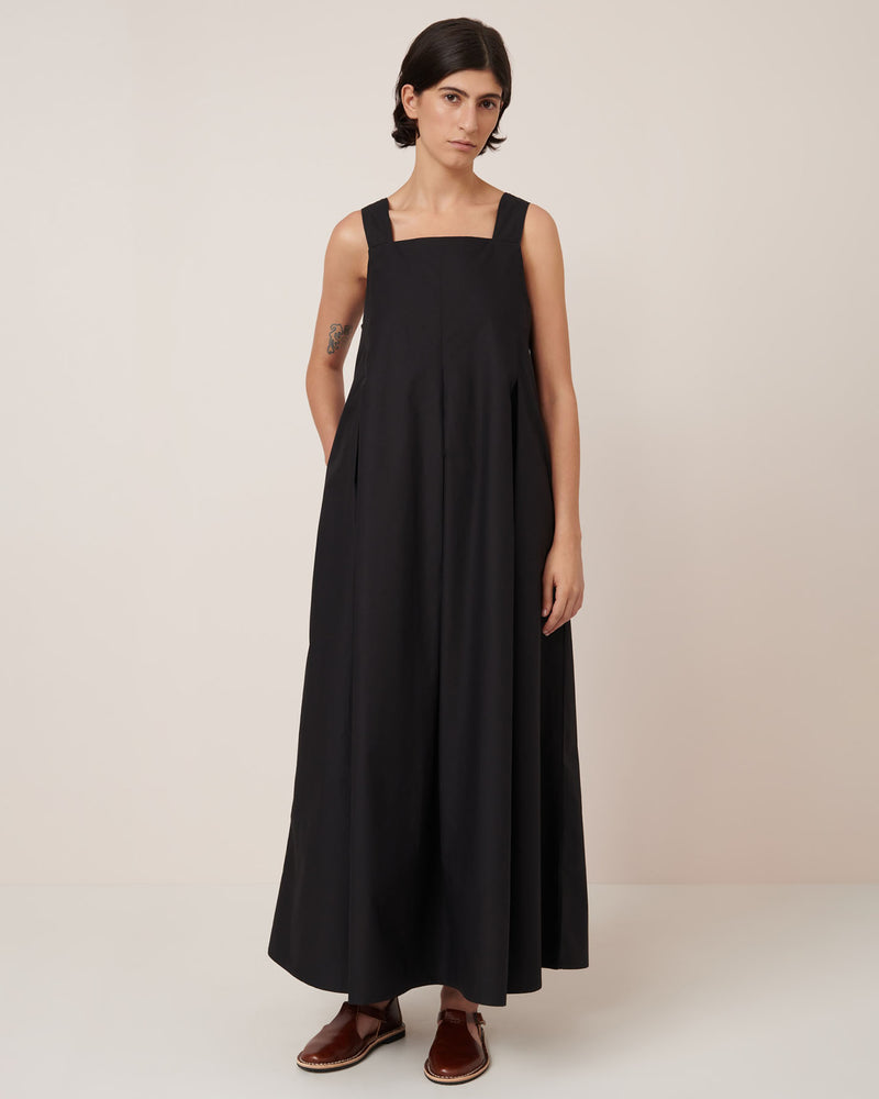 A person with short dark hair wears the Kowtow Study Dress in black, a sleeveless, loose-fitting maxi dress, paired with brown slip-on shoes and stands against a plain light background.