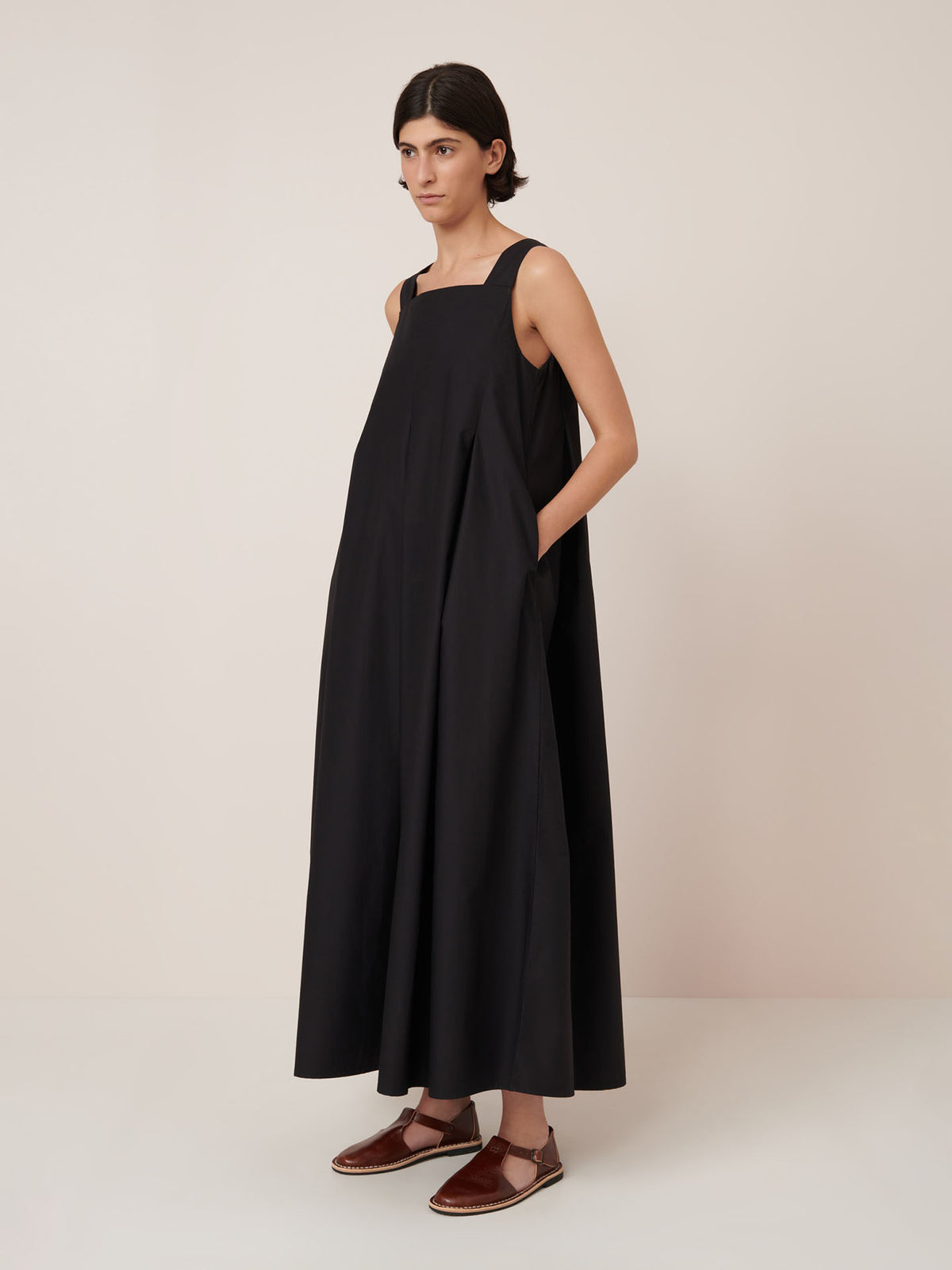 A person stands against a plain background wearing the Kowtow Study Dress in Black, a loose-fitting, sleeveless style paired with brown flat sandals, showcasing its standard true-to-size fit.