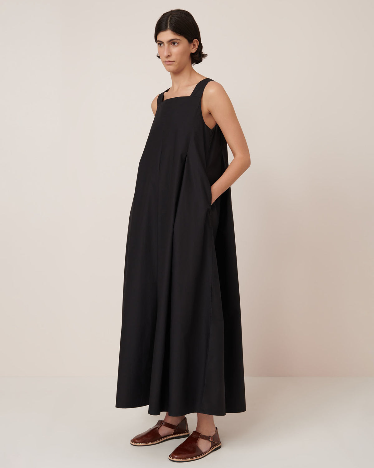 A person stands against a plain background wearing the Kowtow Study Dress in Black, a loose-fitting, sleeveless style paired with brown flat sandals, showcasing its standard true-to-size fit.