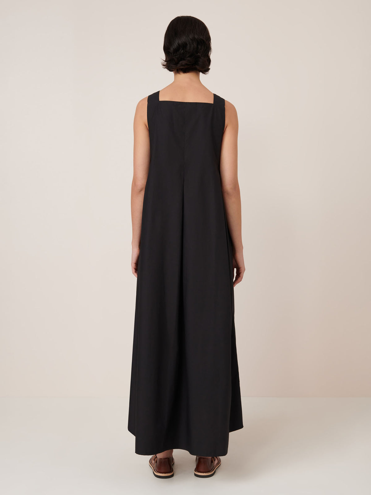 A person stands facing away from the camera against a plain, light background, wearing the Kowtow Study Dress – Black, an ankle-length sleeveless dress with a standard fit for true-to-size comfort. Sandals complete the look.