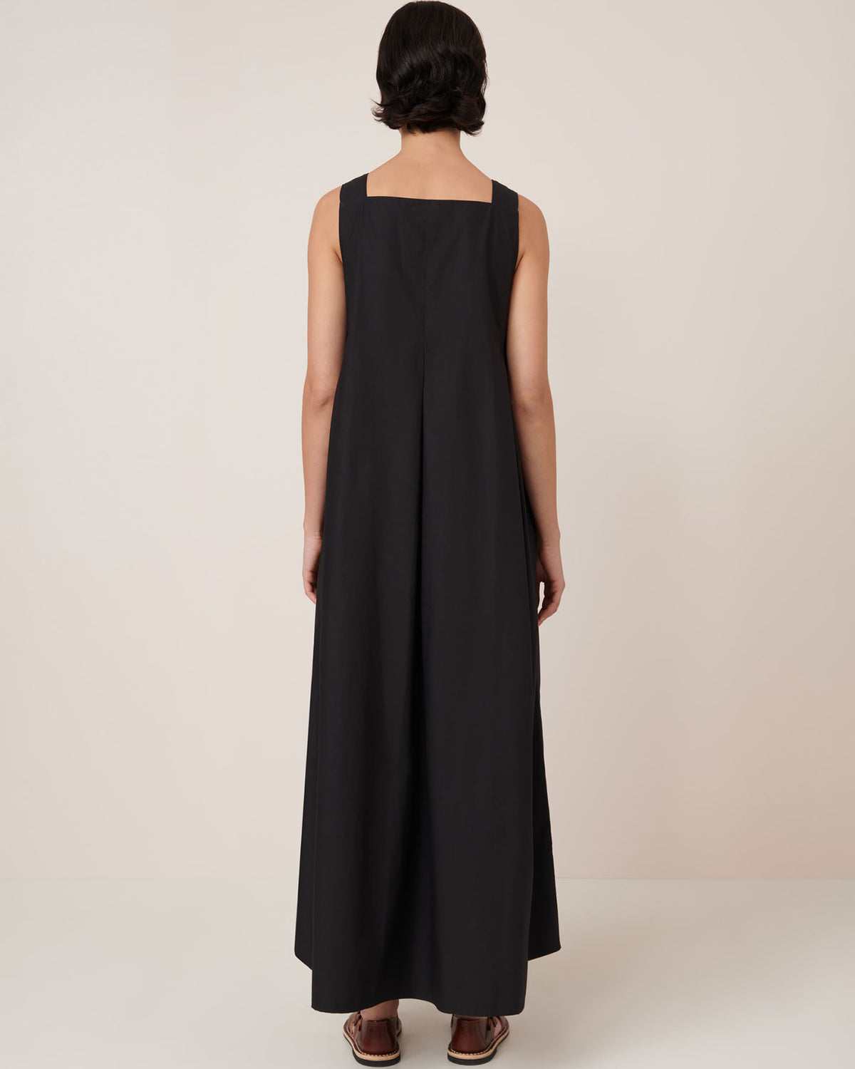 A person stands facing away from the camera against a plain, light background, wearing the Kowtow Study Dress – Black, an ankle-length sleeveless dress with a standard fit for true-to-size comfort. Sandals complete the look.