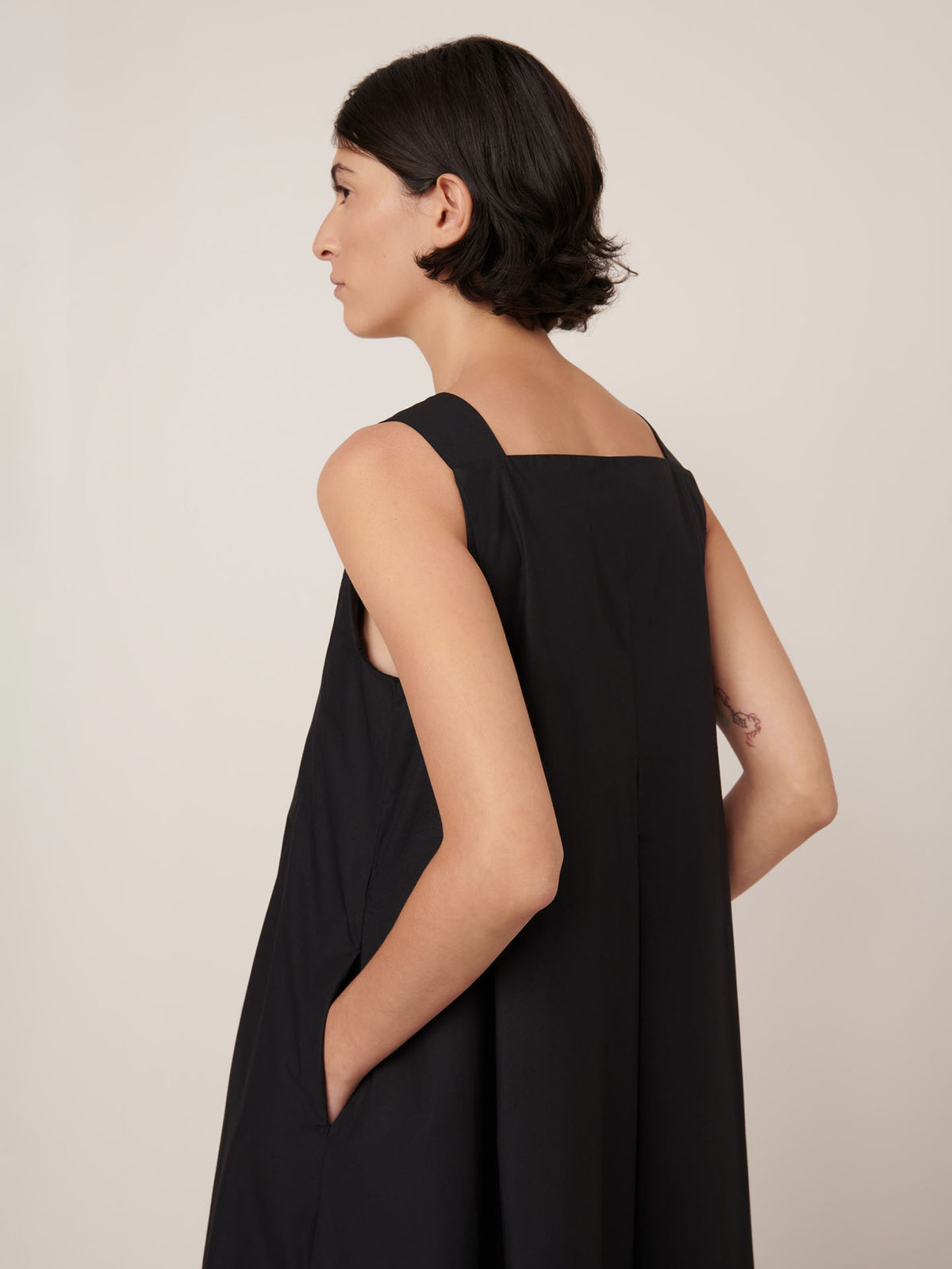 A woman with short dark hair is wearing the Kowtow Study Dress in black—a sleeveless, standard-fit dress—standing with her hands in her pockets and facing left.